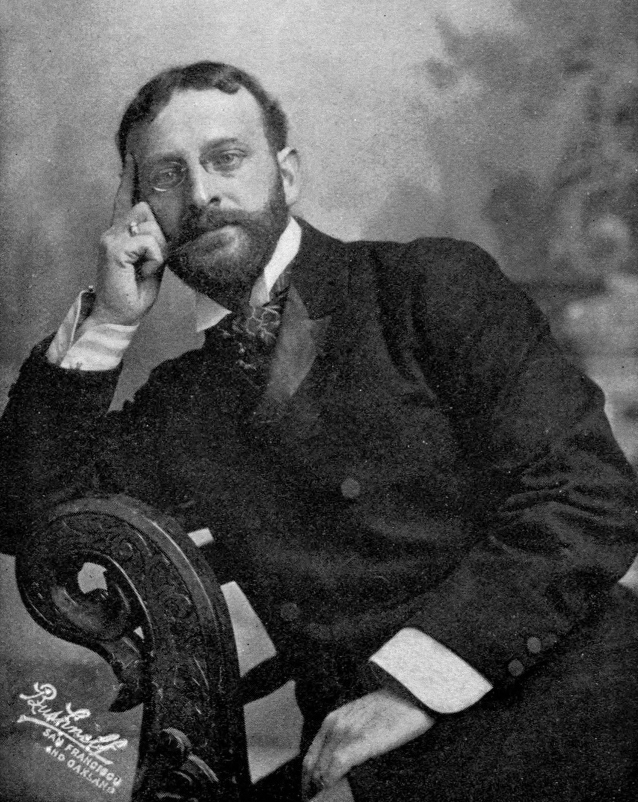 Black-and-white studio portrait of a bearded man in a dark suit wearing pince-nez glasses, seated with one hand raised to his temple.