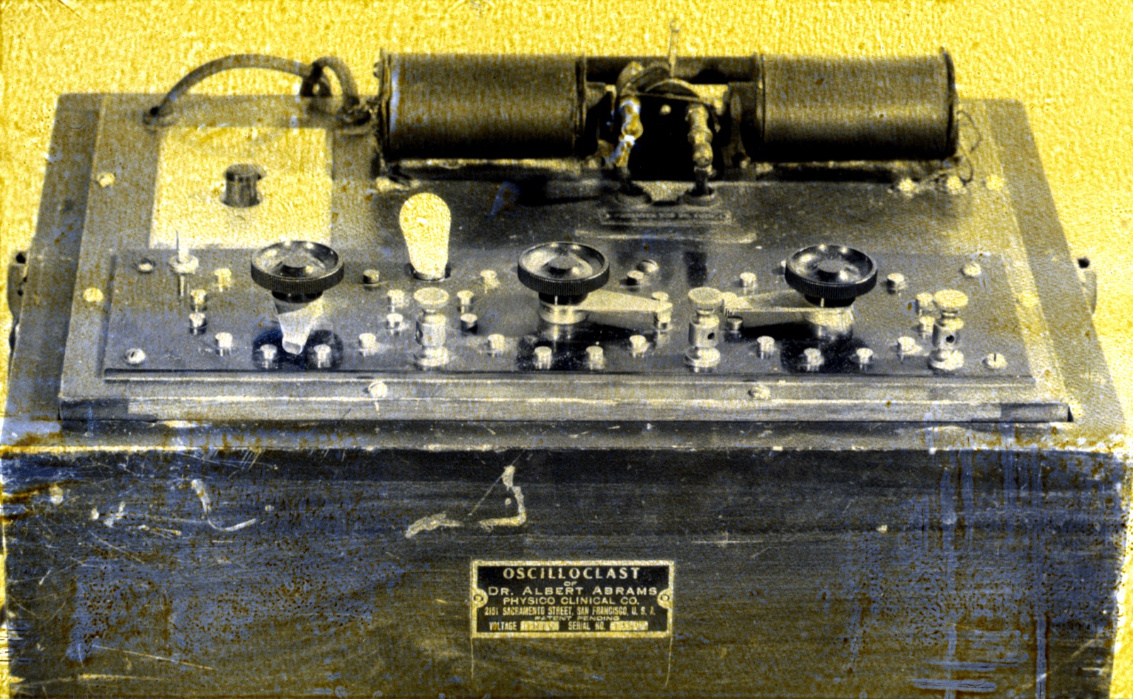 A boxed electrical apparatus with knobs, dials, and a coil mounted on top; a metal nameplate on the front identifies it as an Oscilloclast made by Dr. Albert Abrams's Physico Clinical Co. of San Francisco.