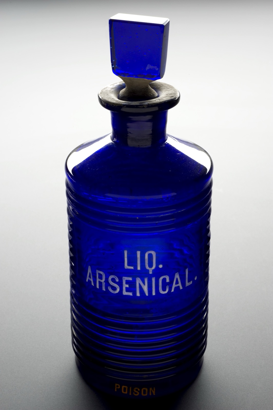 Blue ridged glass pharmacy bottle with a square stopper, labelled 'Liq. Arsenical' and 'Poison'.