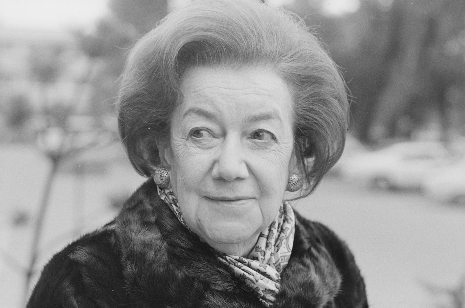 Black-and-white outdoor head-and-shoulders portrait of an older woman with gray hair swept up, round textured earrings, a patterned scarf at the neck, and a dark fur coat, smiling faintly, with a blurred street behind her.