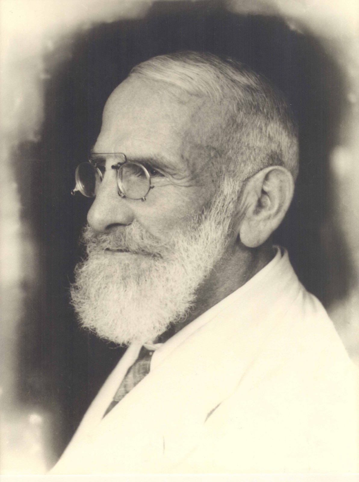 Black-and-white profile portrait of an elderly white-bearded man wearing round spectacles and a light jacket.