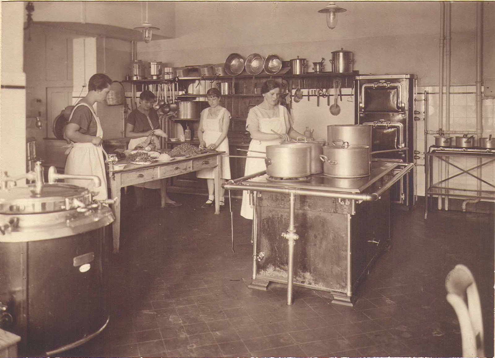 A 1920s institutional kitchen with large stockpots and a central cooking range; several aproned staff work at a preparation table.