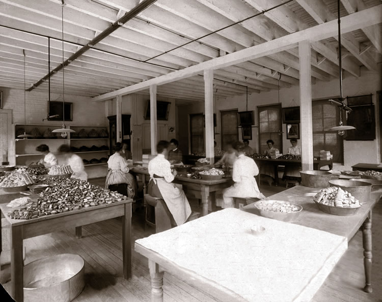 Black-and-white photograph of a factory packing room with several women at long tables filling round tins with pills, from open bowls and boxes of loose pills, under a wood-beamed ceiling