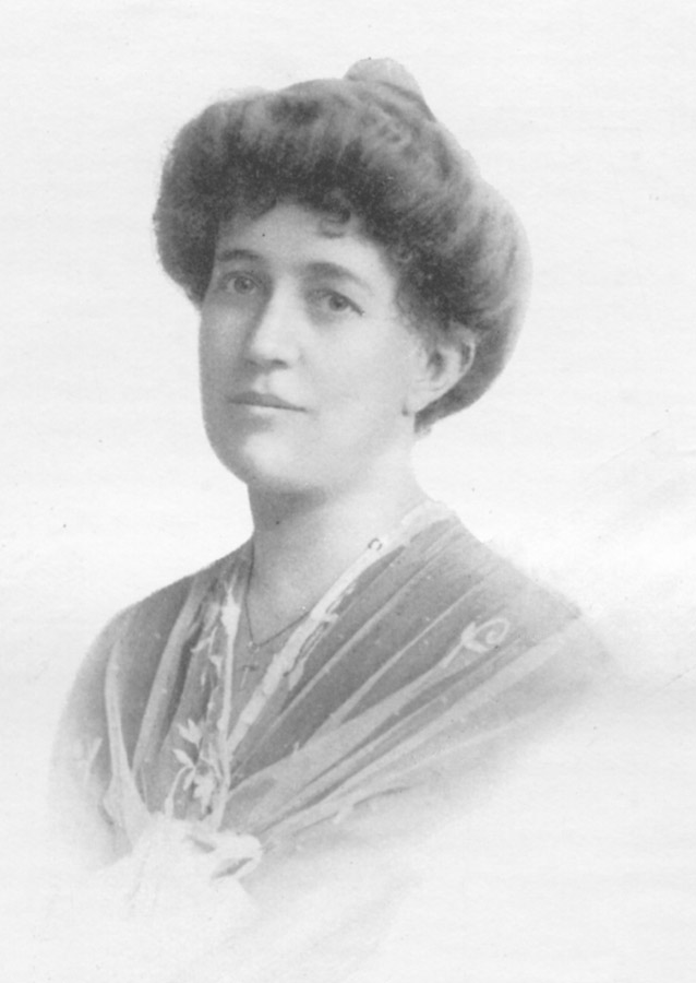 Black-and-white studio portrait of a woman with curled upswept hair, wearing a patterned wrap over a light dress with a small pendant necklace, looking slightly off-camera.