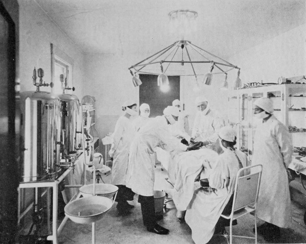 A hospital operating room with a masked, gowned surgical team gathered around a draped patient under an overhead lamp array.
