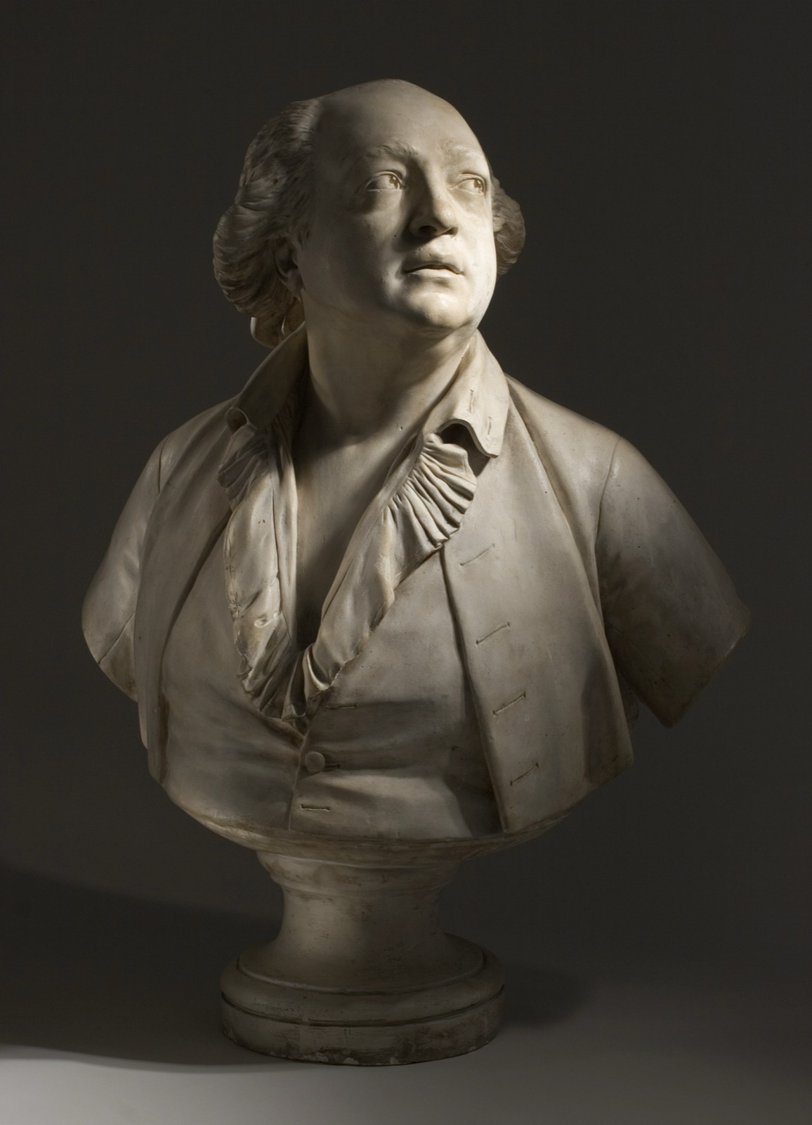 A white marble portrait bust of a heavy-featured man wearing an open eighteenth-century coat and ruffled shirt, his head turned and tilted slightly upward, mounted on a round socle.