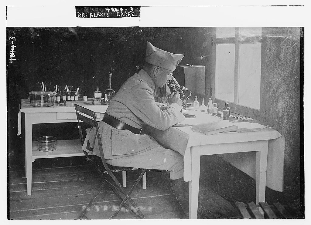 Black-and-white photograph of Dr. Alexis Carrel in military uniform seated at a laboratory microscope, with chemical bottles and glassware on benches around him. Printed caption reads 'DR. ALEXIS CARREL.'