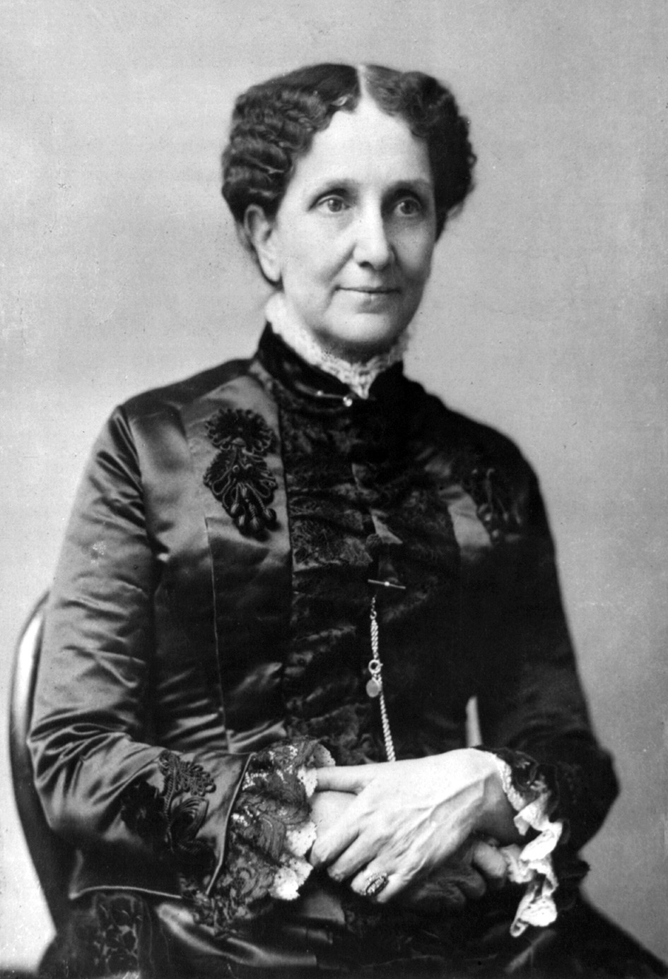Black-and-white studio portrait of an older woman seated, wearing a dark satin dress with lace cuffs, hands folded in her lap.