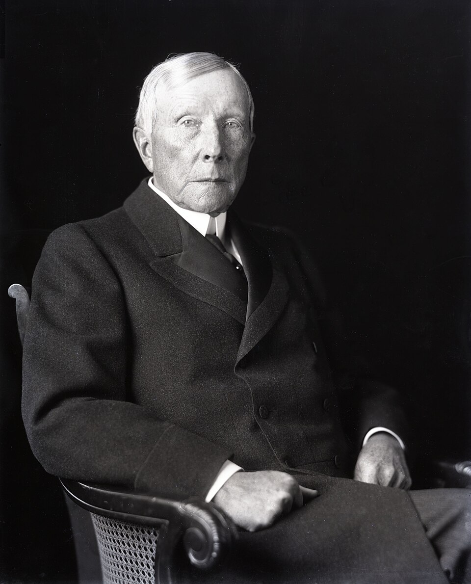 Black-and-white seated portrait photograph of John D. Rockefeller, an older man in a dark suit and white collar, facing the camera from an armchair.