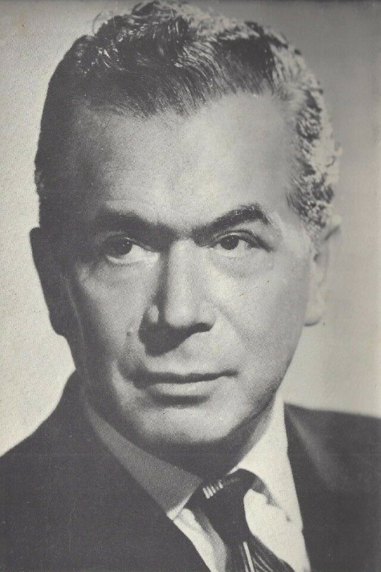 Black-and-white studio portrait photograph of an older man with swept-back grey hair, wearing a dark suit and striped tie.
