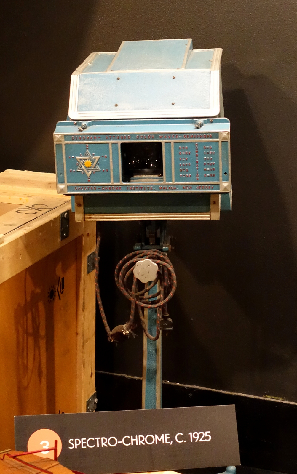 A teal-blue metal box-shaped device on a stand, labeled Dinshah-Attuned Color Waves-Semaphore and Spectro-Chrome Institute, Malaga, New Jersey, with a central dark lamp aperture, a six-pointed star wheel diagram with color-code letters, a Combination code chart, and a coiled power cord wound around the stand.