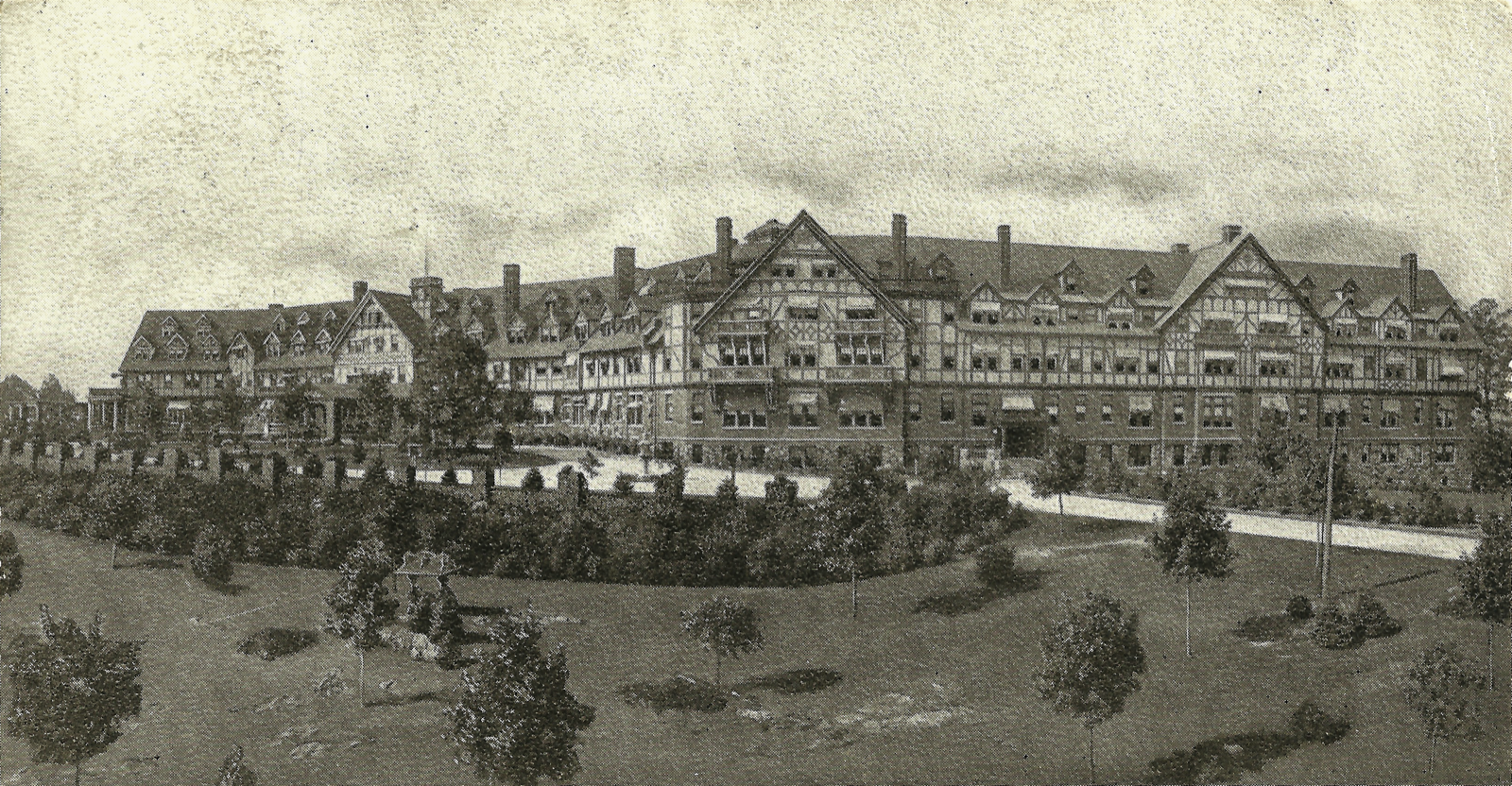 Black-and-white photograph of a large multi-winged Tudor-style resort hotel with many gables and chimneys, set on landscaped grounds with young trees and hedged paths.