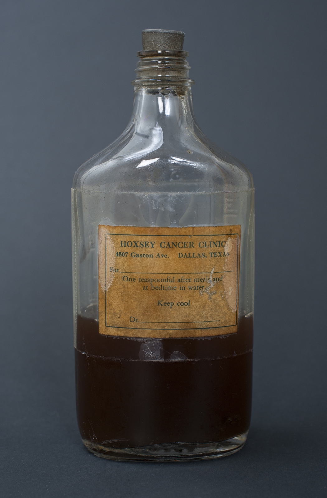 A clear glass bottle roughly two-thirds full of dark brown liquid, with a cork stopper and a paper label reading Hoxsey Cancer Clinic, 4507 Gaston Avenue, Dallas, Texas, with dosing instructions.