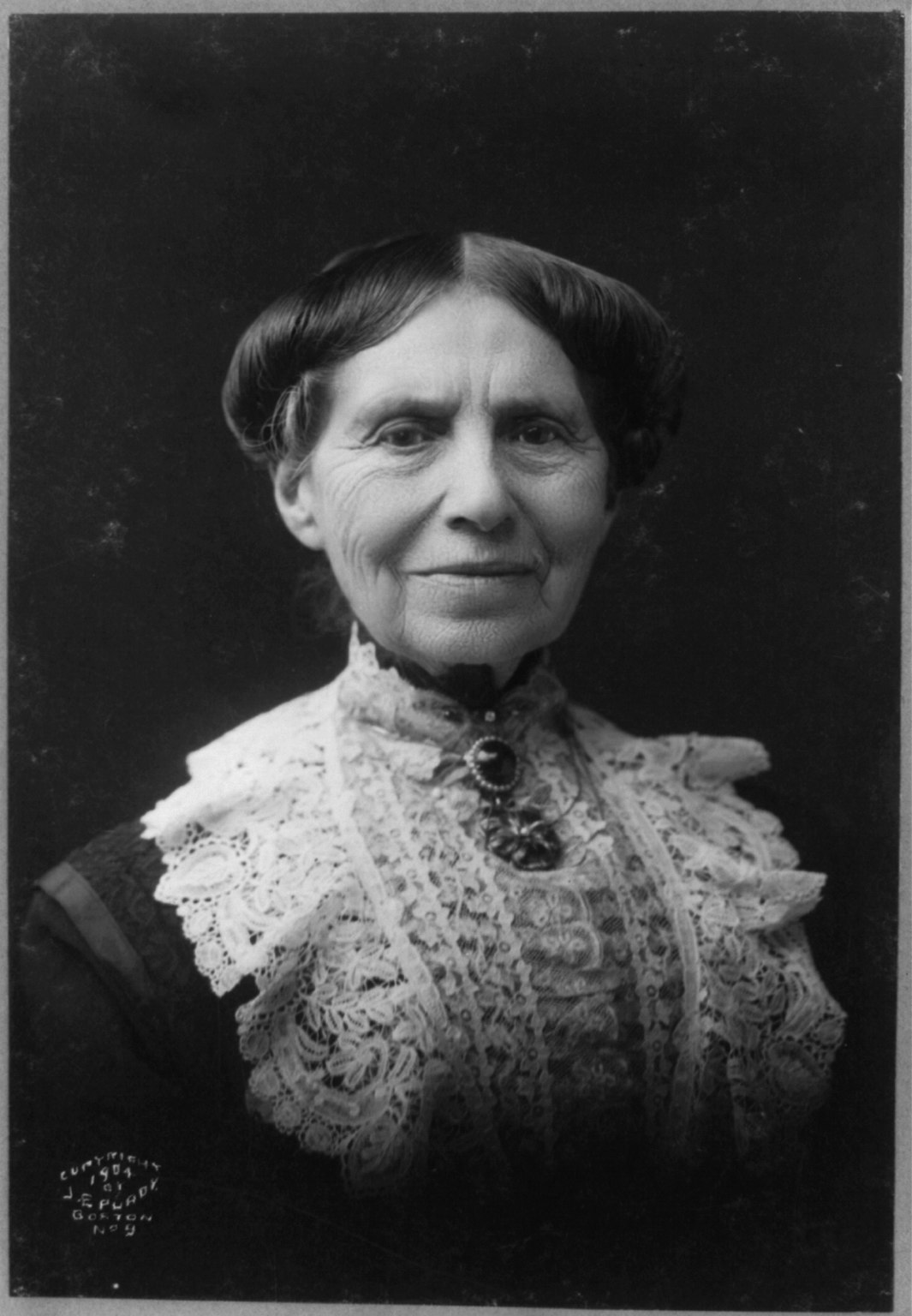 Black-and-white head-and-shoulders studio photograph of an older woman with dark hair pulled back, facing the camera, wearing a dark high-collared dress.