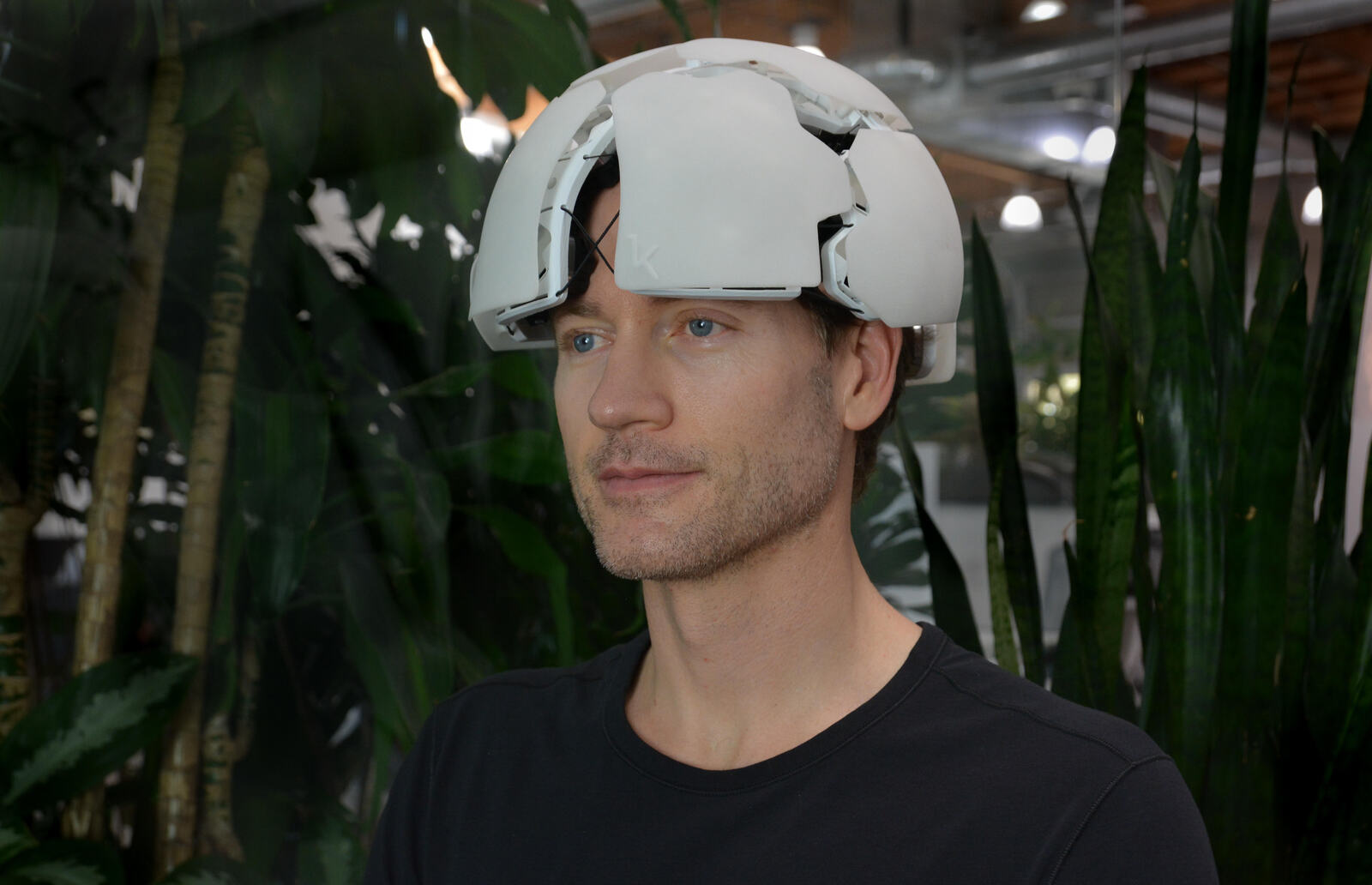 A man in a black t-shirt wearing a white Kernel Flow brain-imaging headset on his head, photographed indoors among green potted plants.