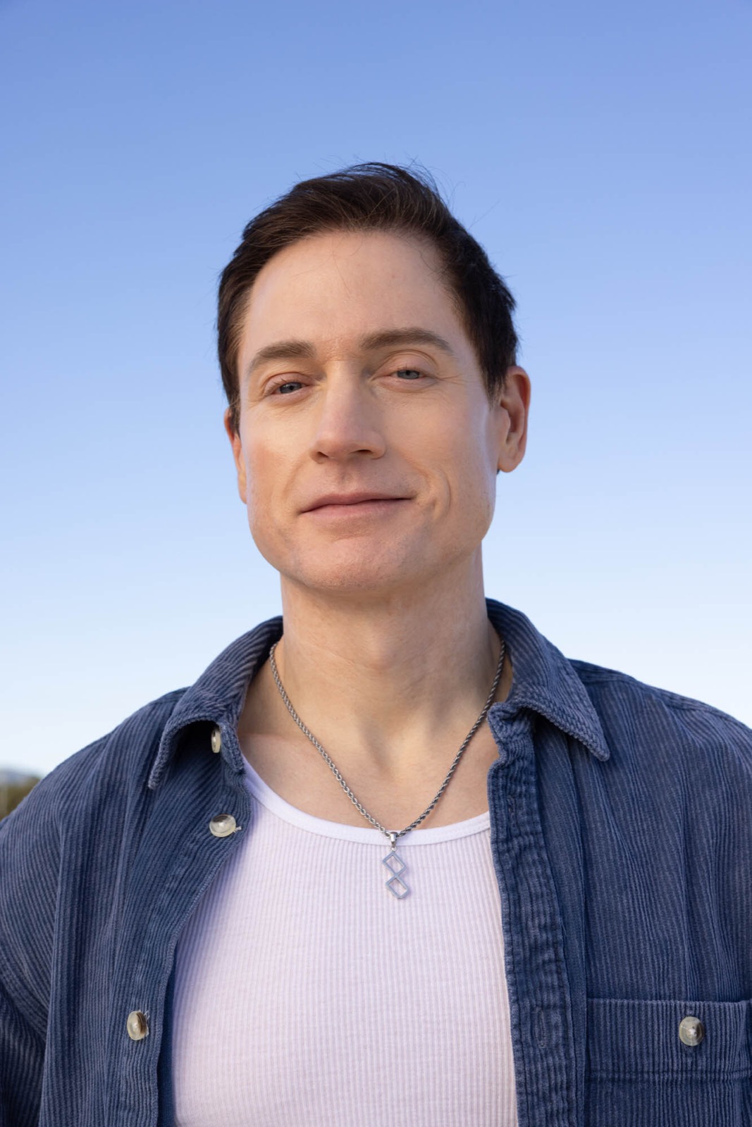 Head-and-shoulders portrait of a man with short dark hair, wearing an open blue corduroy overshirt over a white top and a silver pendant necklace, against a clear blue sky.