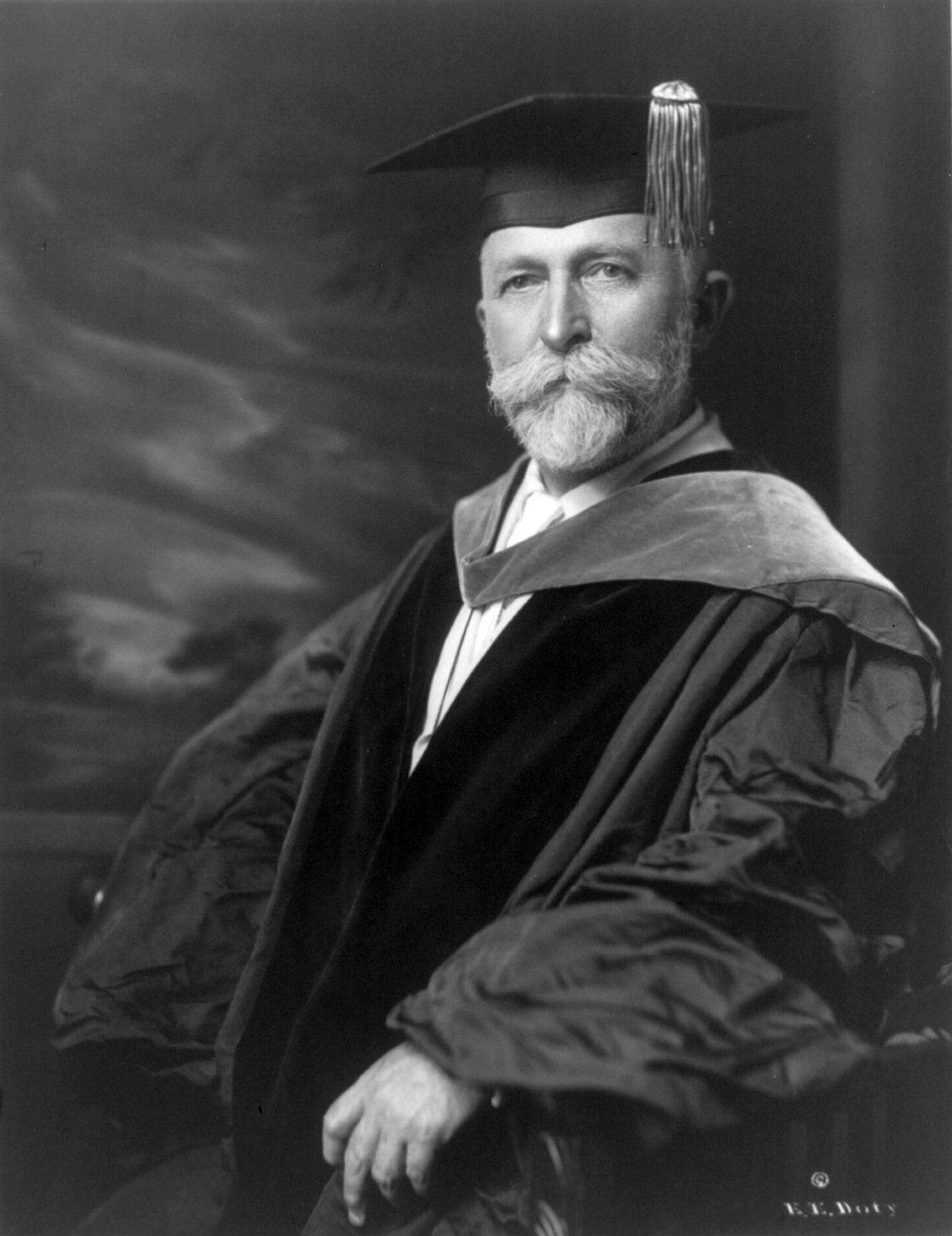Studio portrait of an older bearded man in academic cap and gown, seated against a painted backdrop.