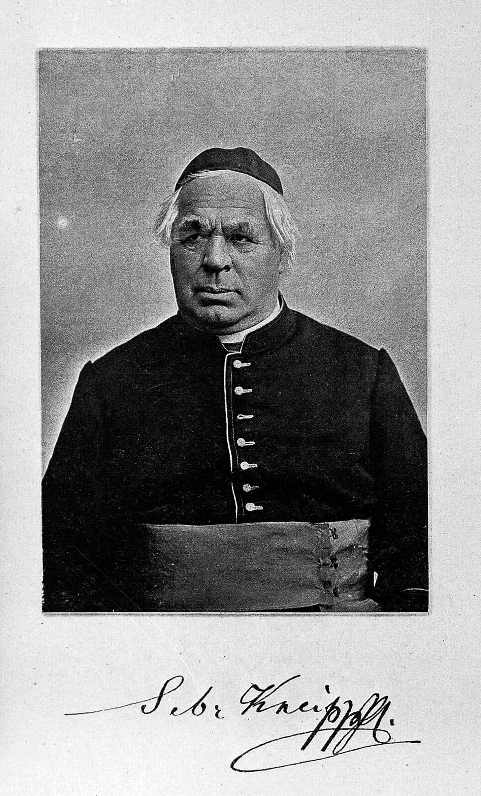 Studio portrait of an elderly man with white hair, wearing a black skullcap and clerical cassock with a white collar, with a cursive signature beneath the image.