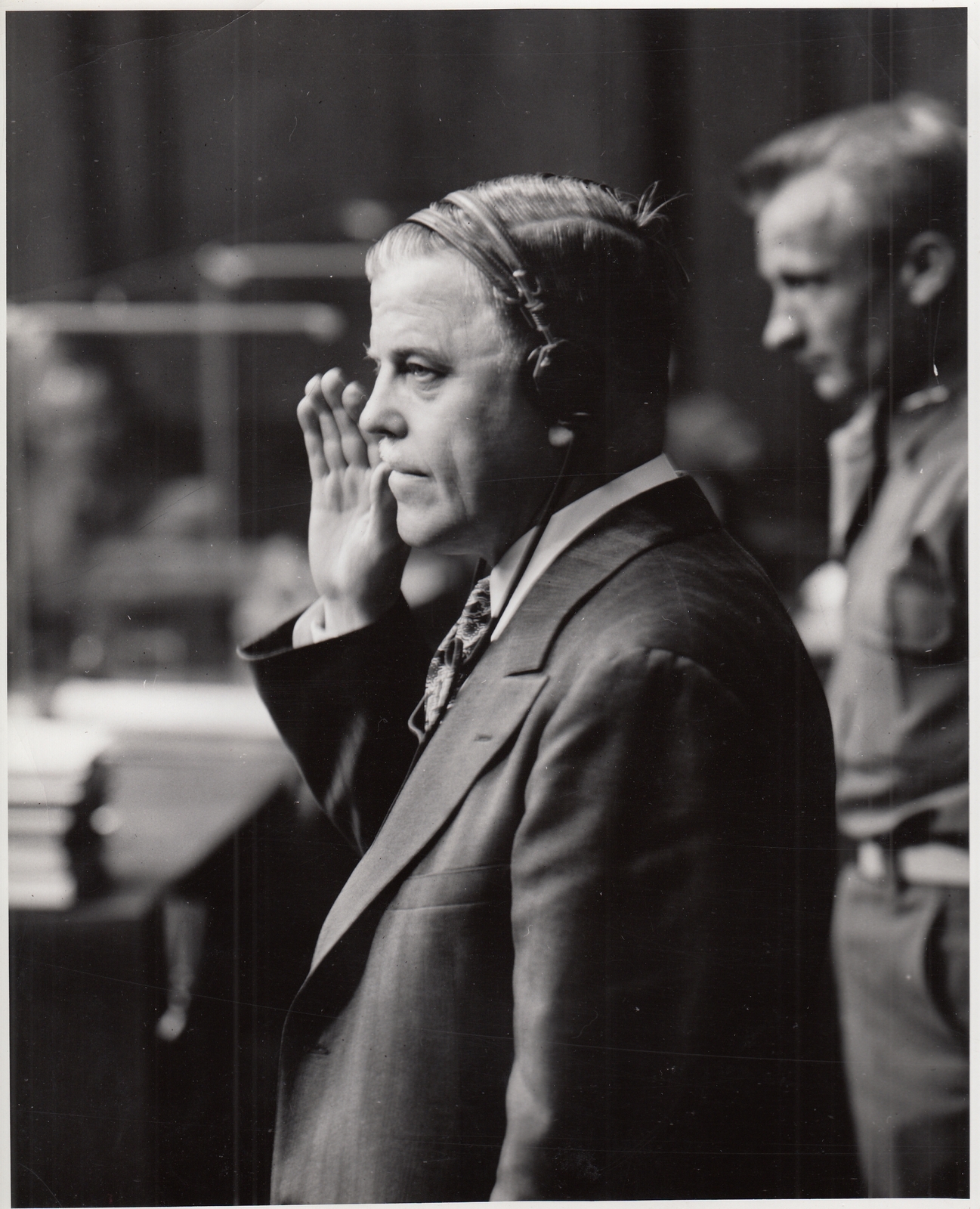Black-and-white photograph of a middle-aged man in a suit standing with his right hand raised, wearing translation headphones, in a formal tribunal setting with a uniformed official behind him.