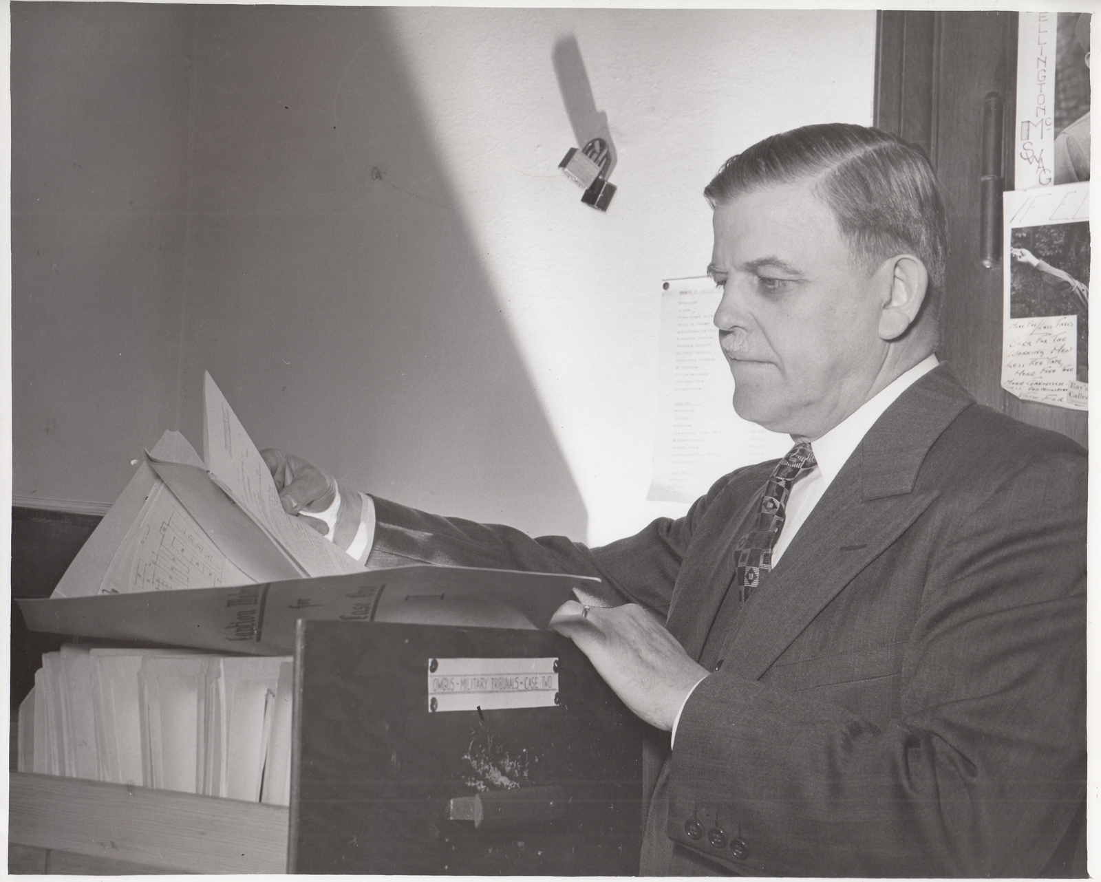 Black-and-white photograph of a man in a suit standing in profile at a lectern or document stand, reviewing papers, with an OMGUS Military Tribunals case label visible on the stand.