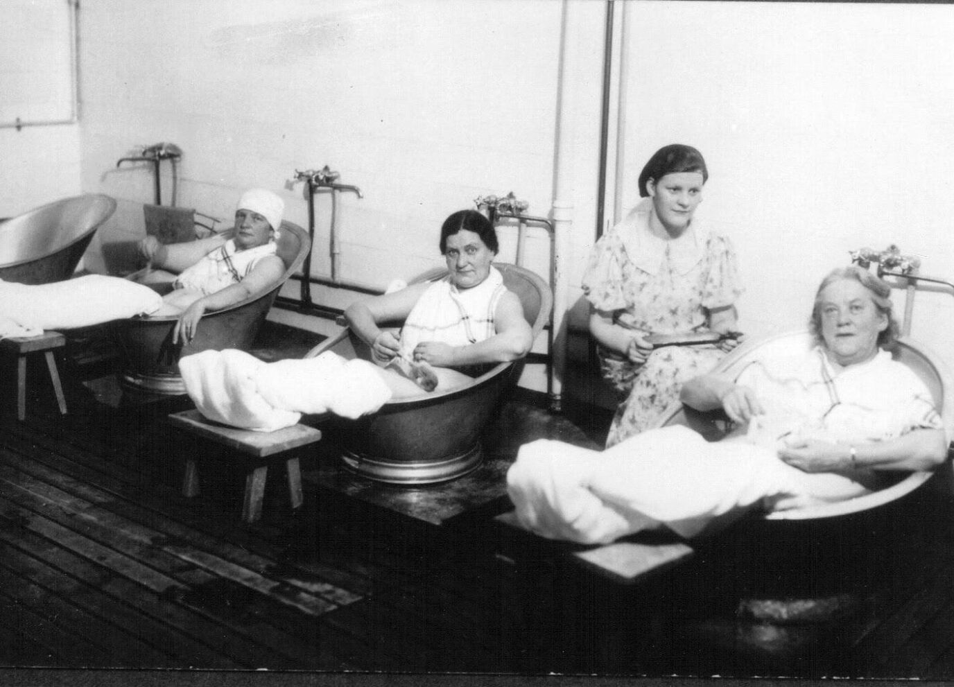 Interior photograph of four women seated in individual metal bathtubs in a plain treatment room, with a young woman in a patterned dress standing among them.