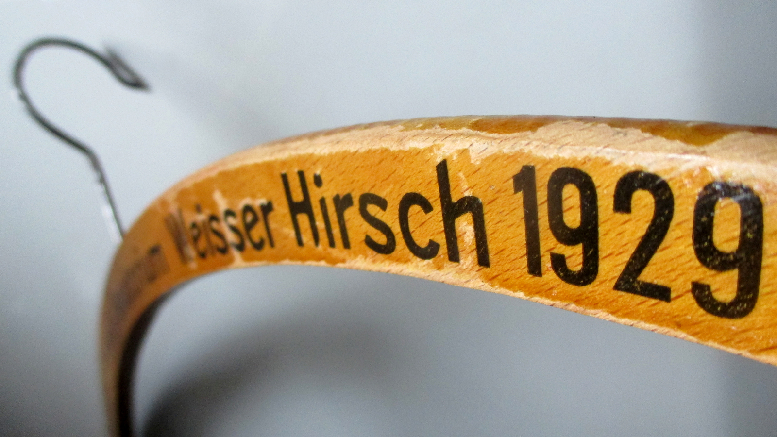Extreme close-up of a curved wooden clothes-hanger arm with a metal hook, printed with partly legible black text ending '...Weisser Hirsch 1929'.