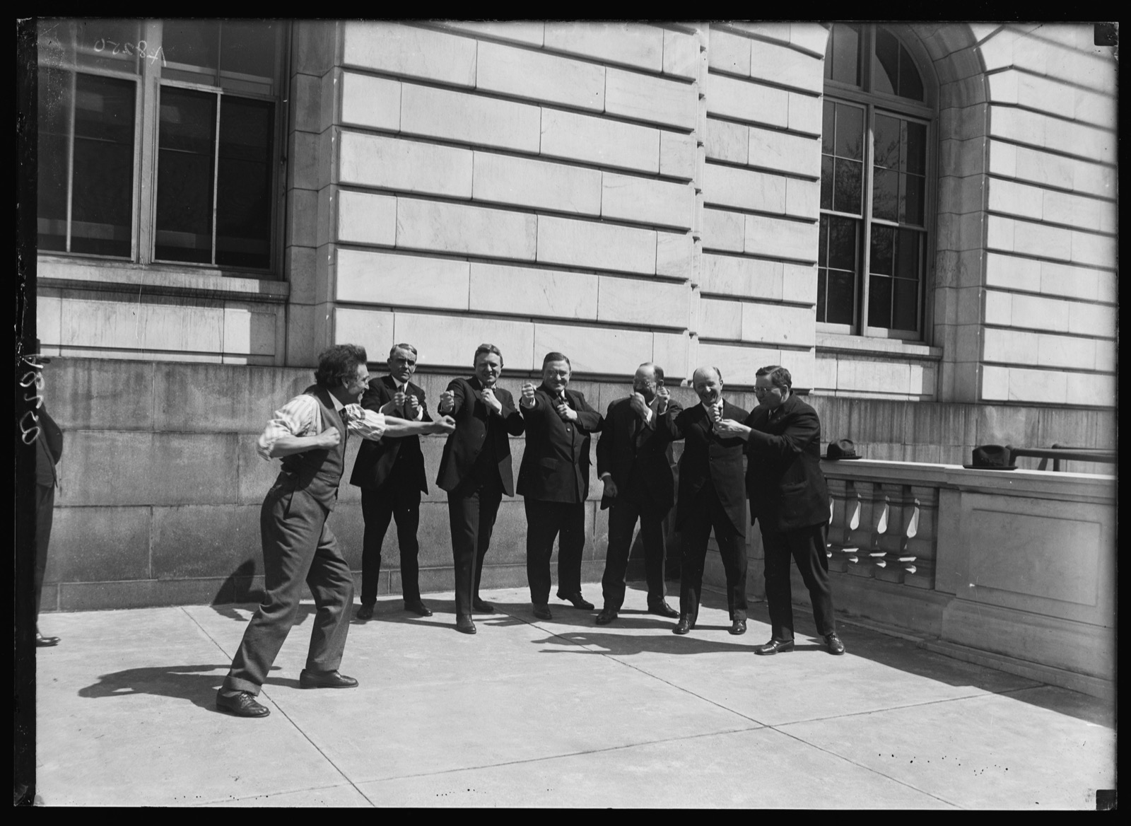 Black-and-white outdoor photograph of a line of men in suits performing a chest-level arm exercise, led by a man in shirtsleeves and suspenders at the left, before a stone building.