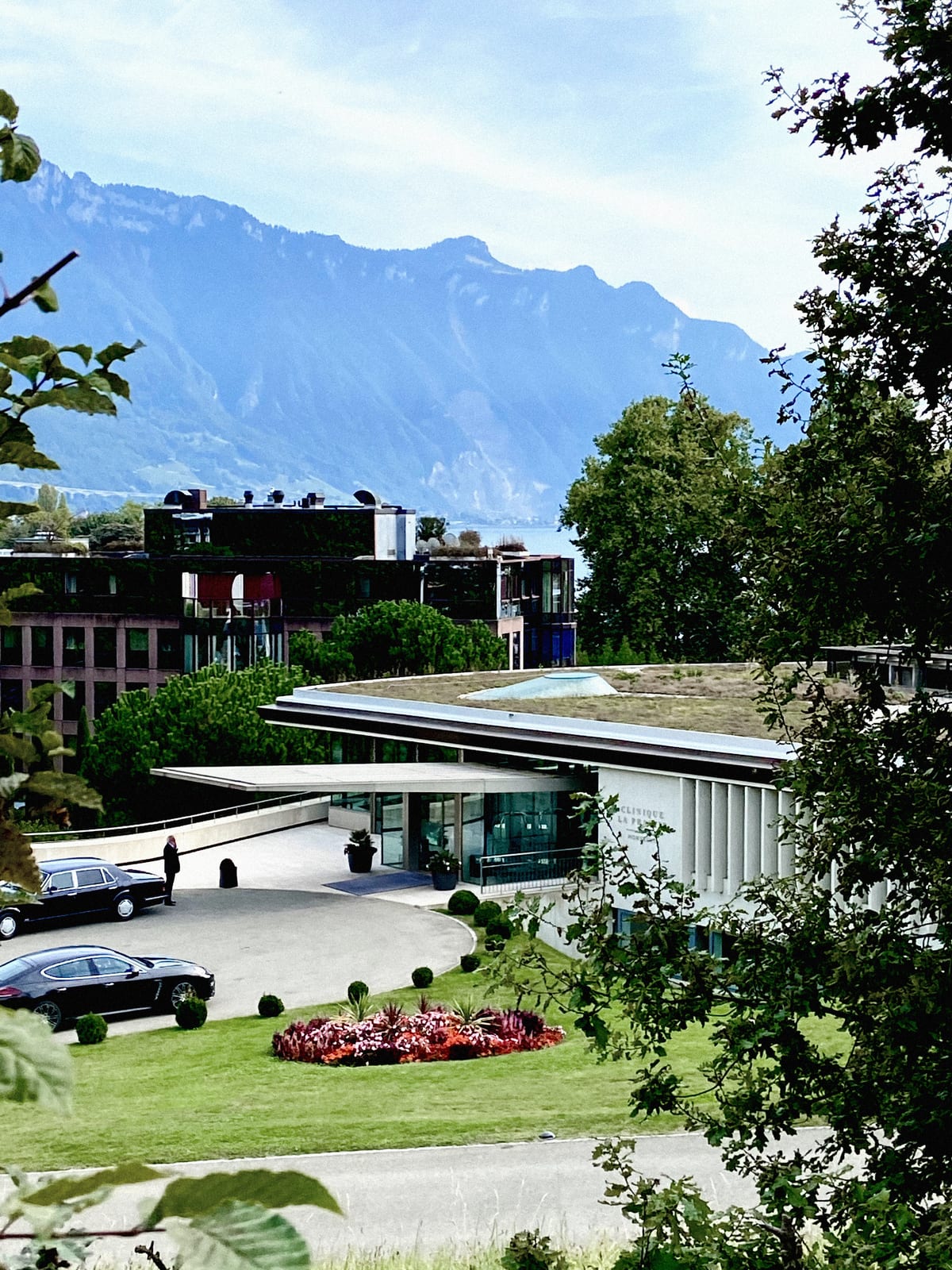 Photograph of the Clinique La Prairie buildings in Montreux, with a road and landscaping in the foreground and mountains in the background.