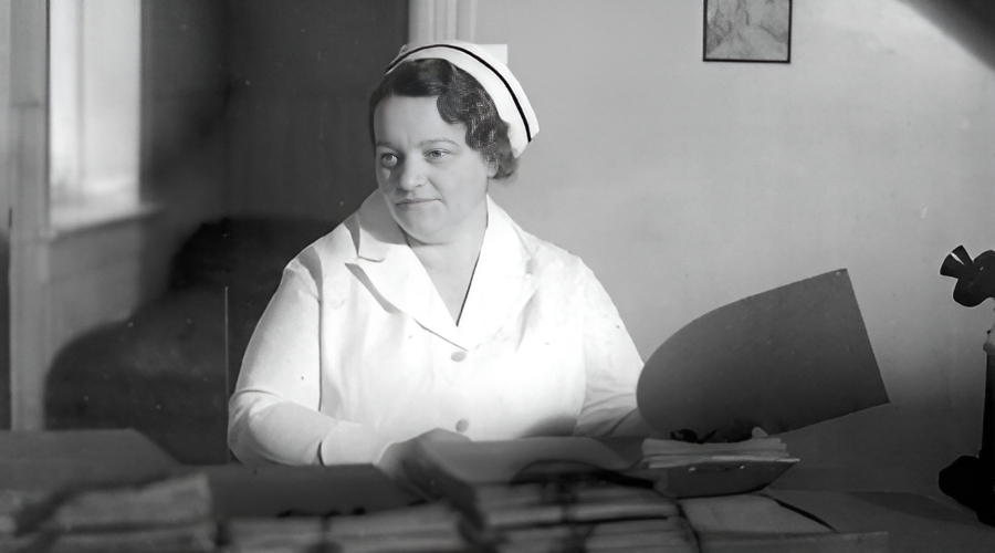 Black-and-white archival photograph of Rene Caisse in a white nurse's uniform and cap, seated at a desk with papers and files.