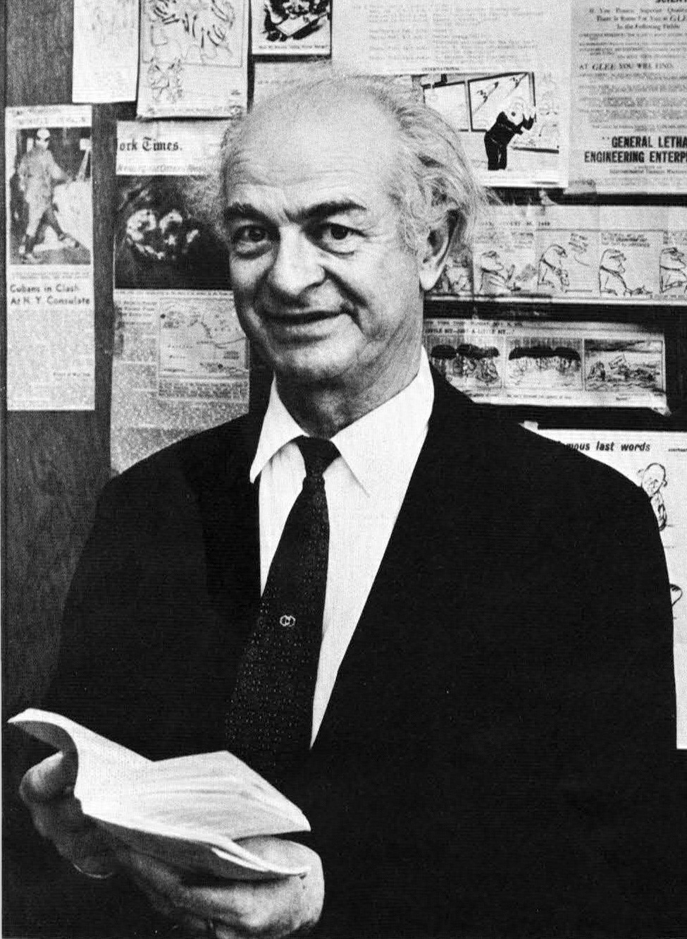 Black-and-white photograph of an older balding man with white hair, wearing a dark suit and tie, smiling slightly while holding an open book, standing in front of a bulletin board covered with newspaper clippings and cartoons.