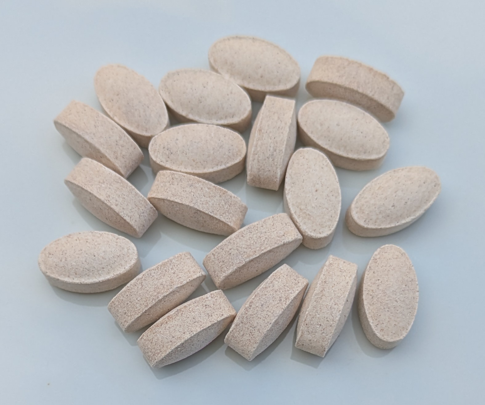 Close-up photograph of about twenty oval, beige, speckled vitamin C tablets scattered on a light gray surface.