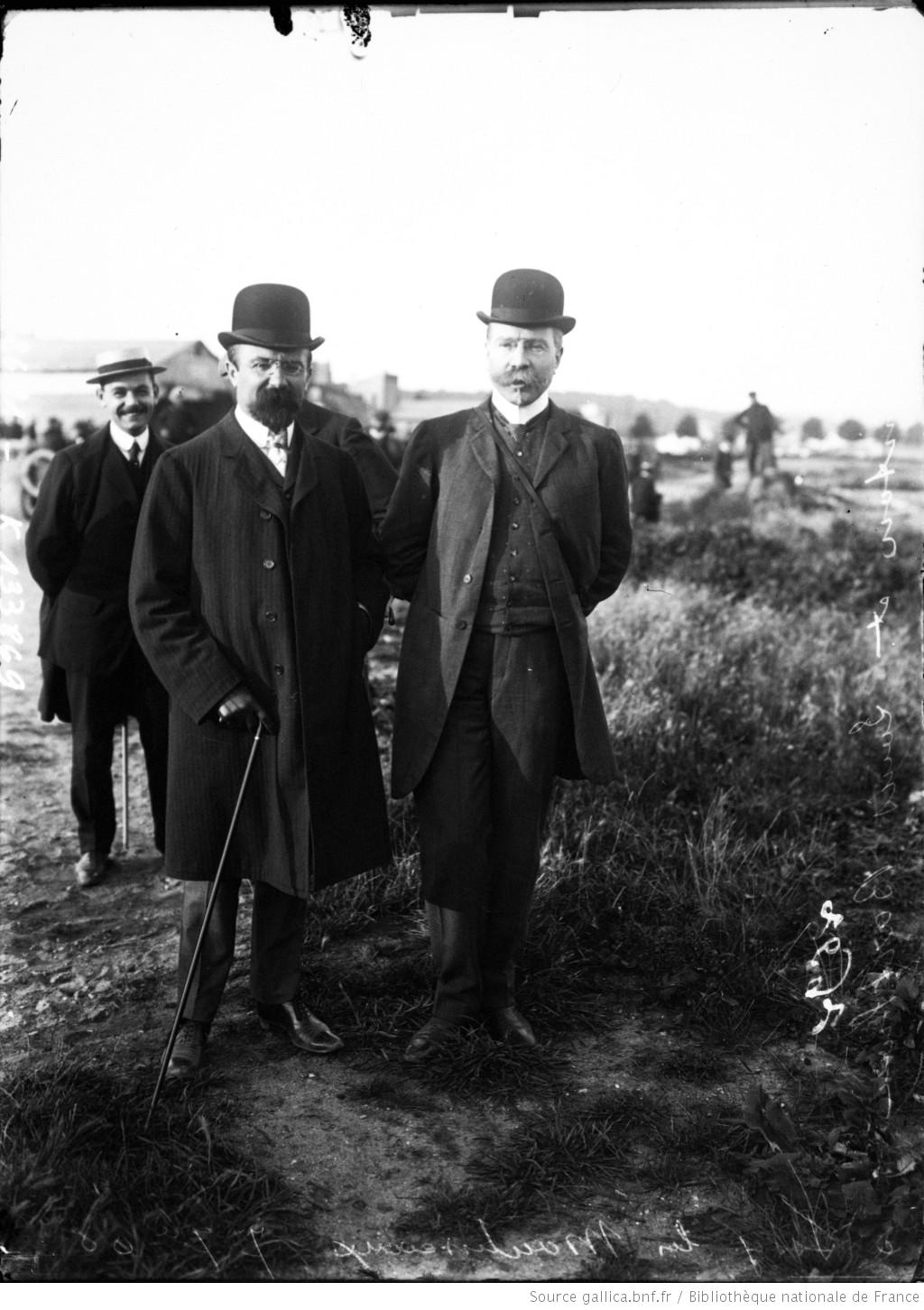 Two men in dark overcoats and bowler hats standing outdoors in an open field, a third man visible in the background