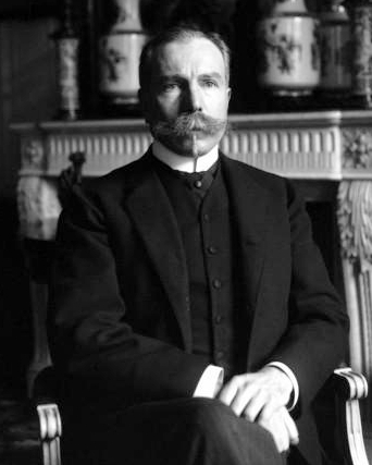 Seated formal portrait of a bearded man in a dark suit against a background of decorative objects