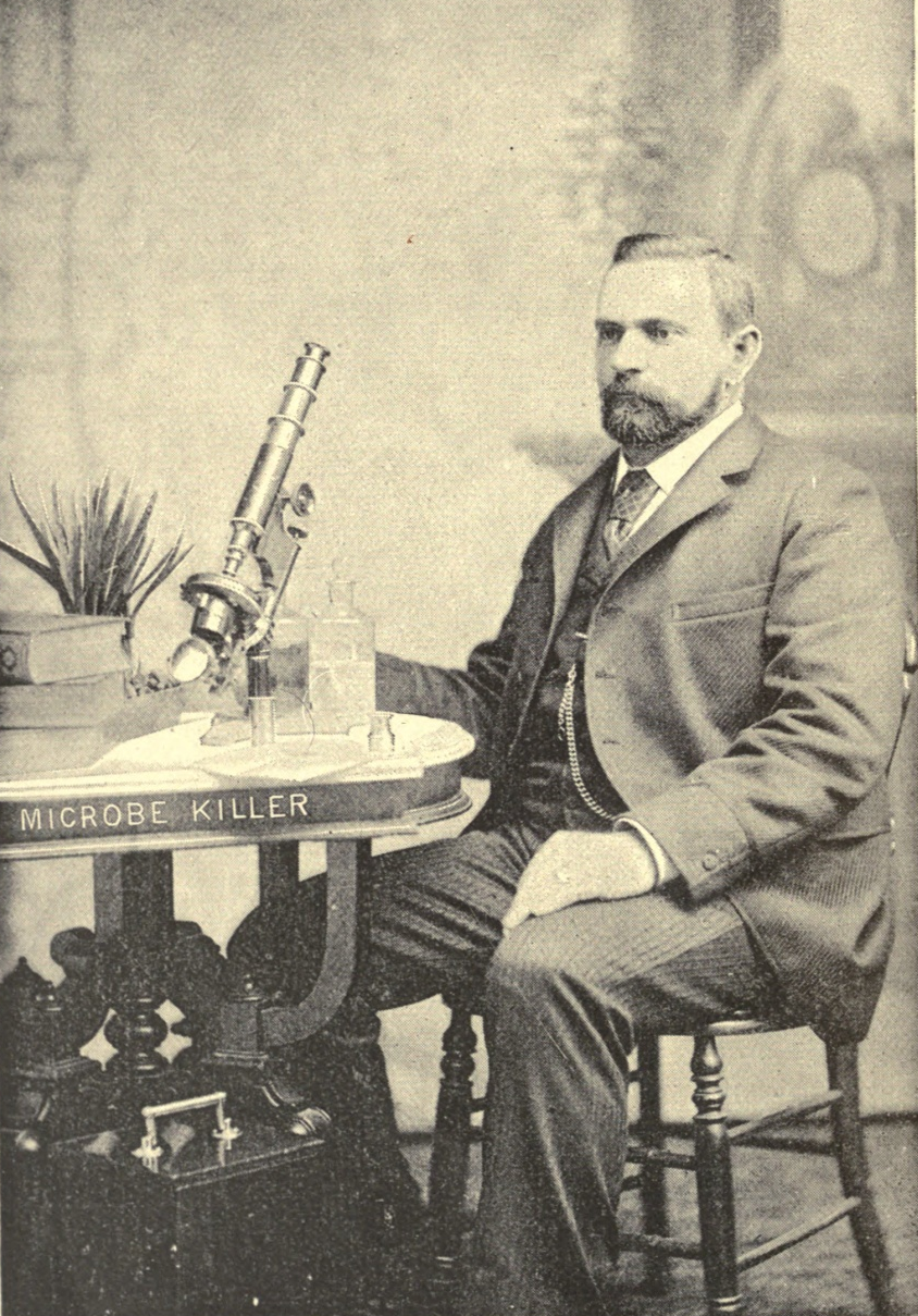 A bearded man in a suit sits beside a small round table holding a microscope and a jug lettered MICROBE KILLER.