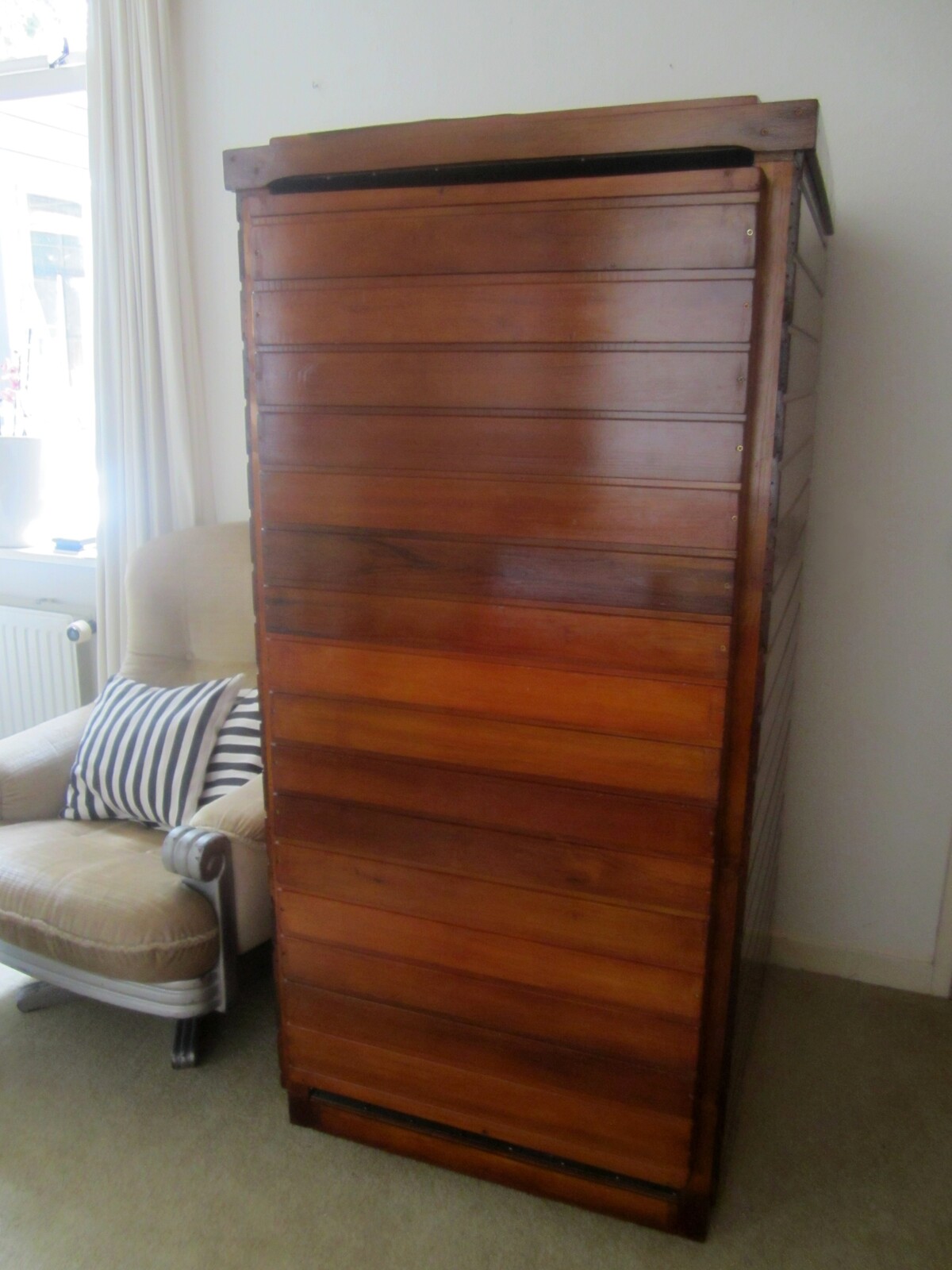 A tall, upright wooden cabinet the height of a person, its front made of horizontal wooden strips in alternating tones, standing in a room beside an armchair.