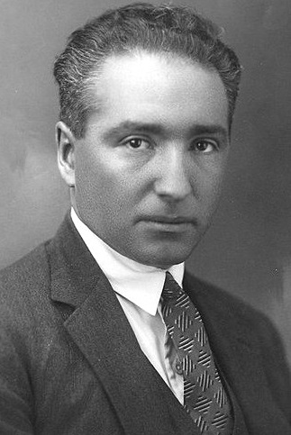 A studio portrait photograph of a man in early middle age with dark, swept-back hair, wearing a dark suit and tie, looking directly at the camera.