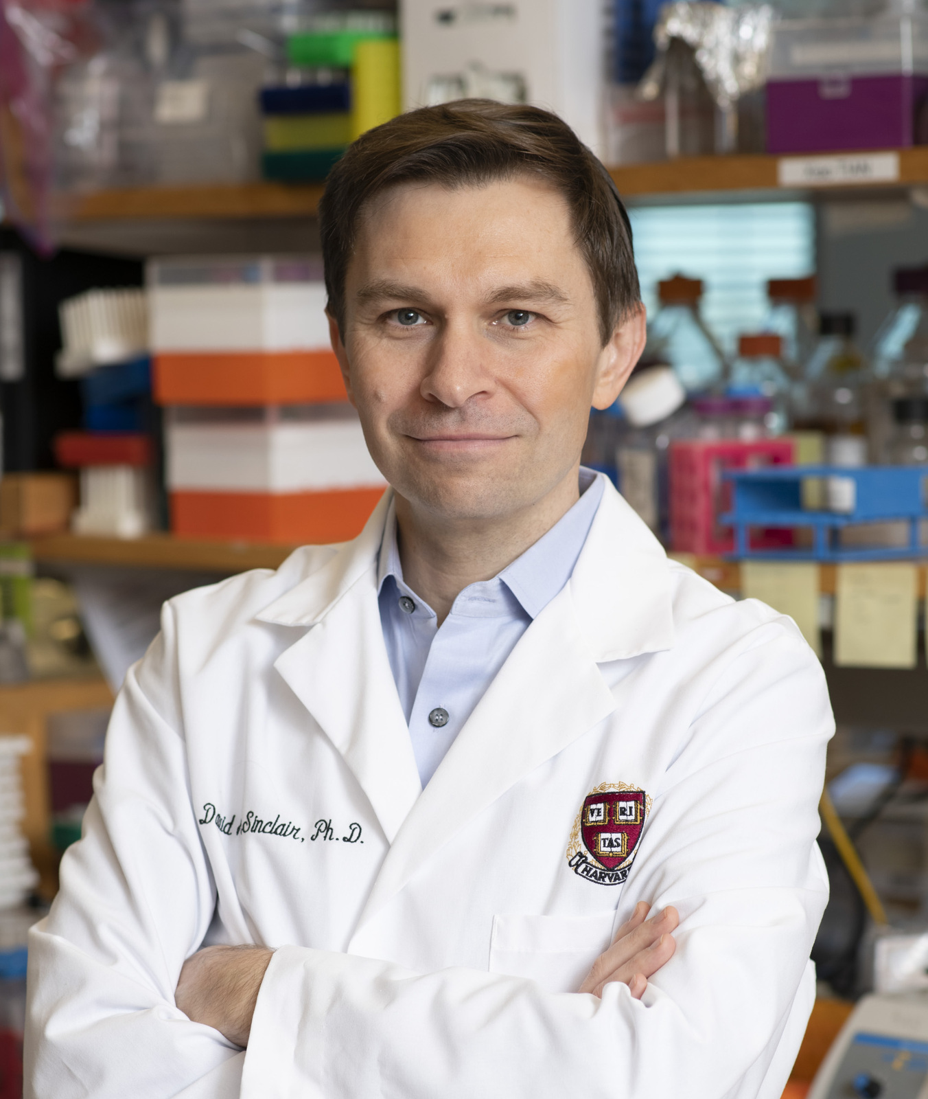 Portrait of a smiling man in a white lab coat embroidered 'David Sinclair, Ph.D.' with a Harvard crest, arms crossed, standing in a laboratory.