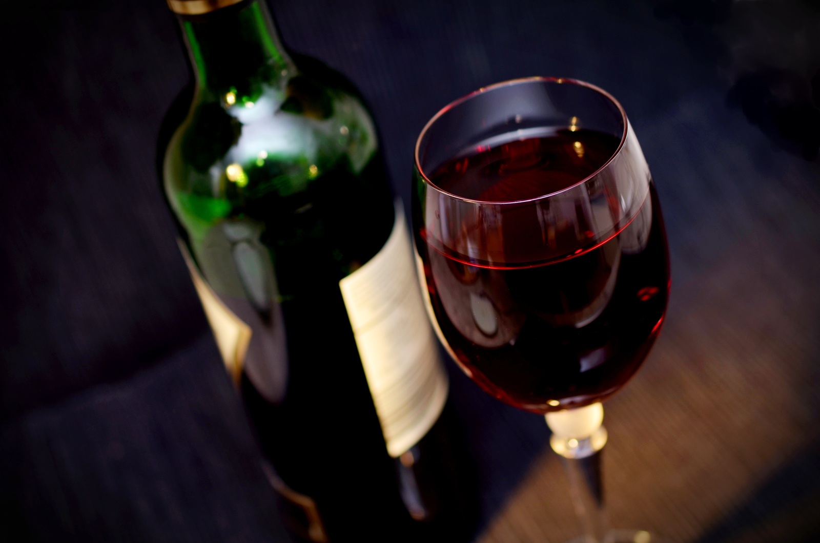 A dark bottle of red wine beside a stemmed glass filled with red wine, on a dark table.
