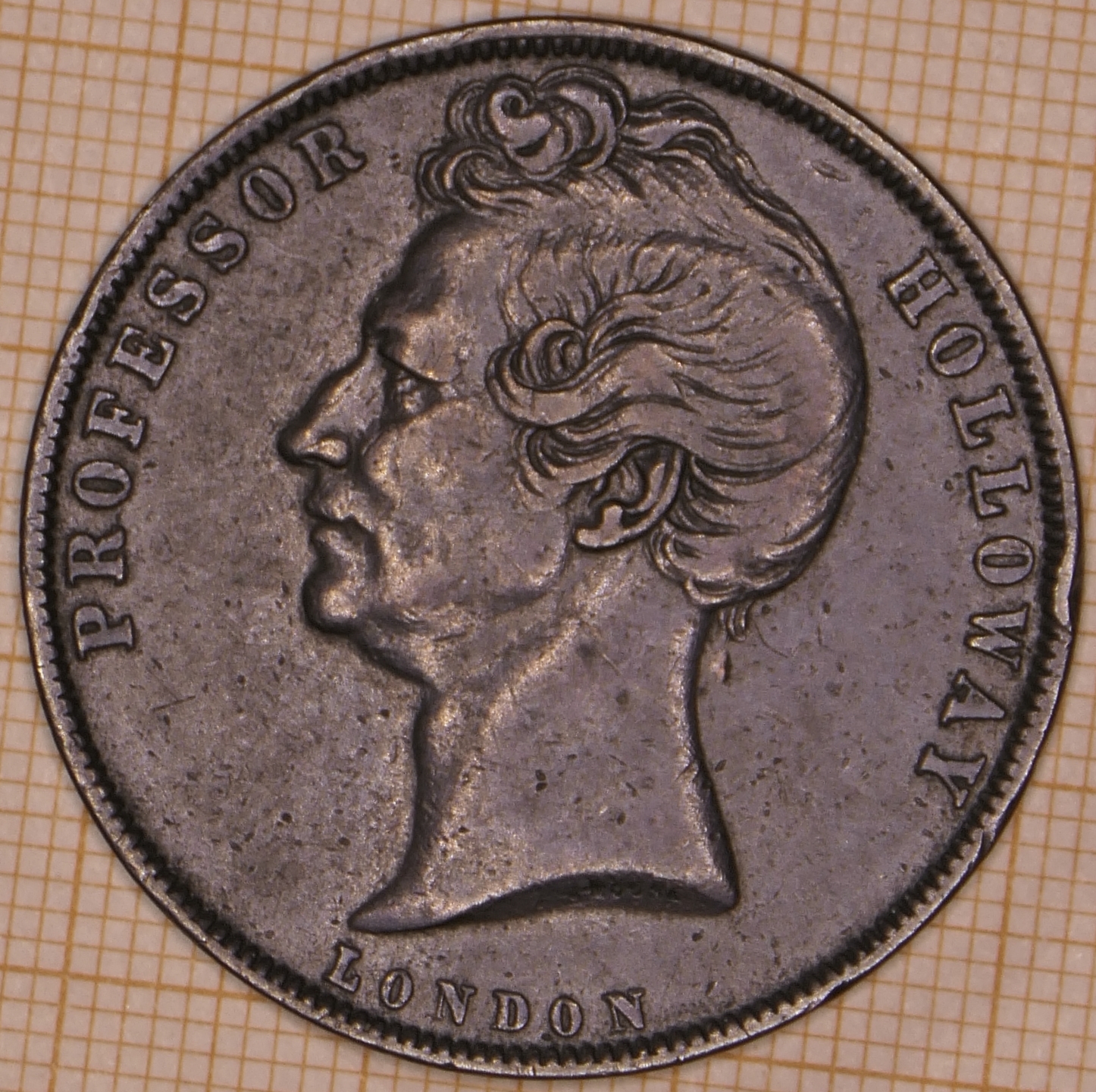 A worn copper coin-like token showing a left-facing male bust with wavy hair, encircled by the words PROFESSOR HOLLOWAY above and LONDON below