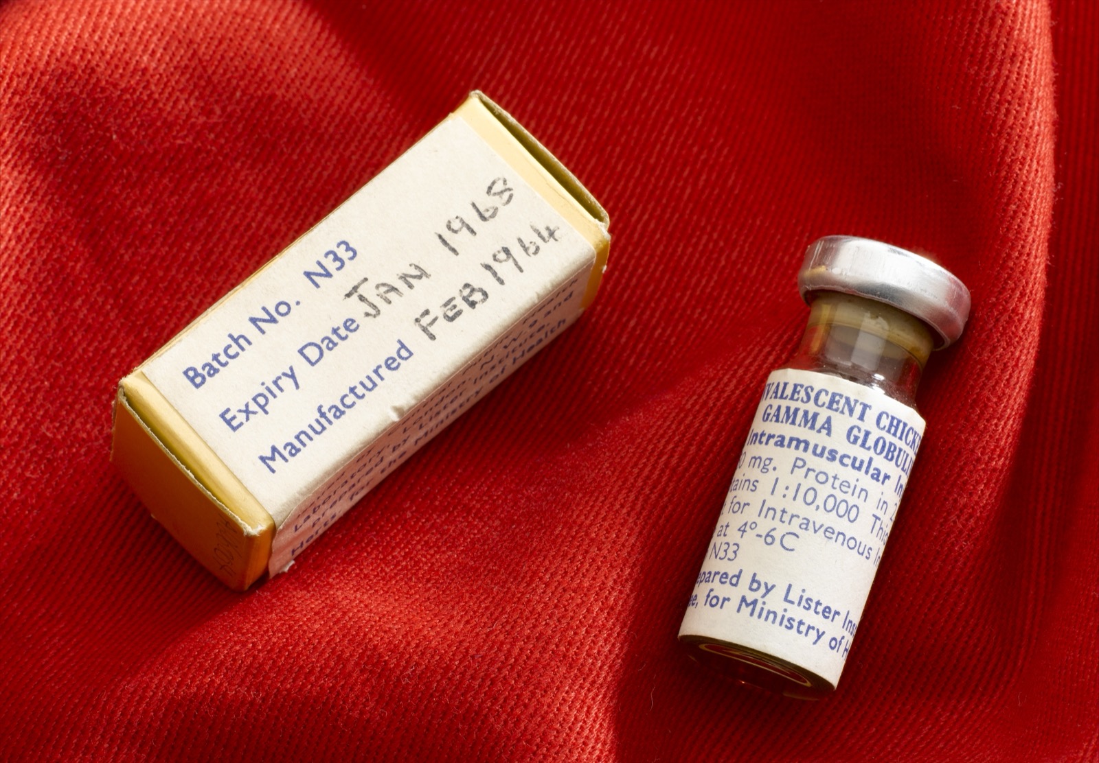 Small vial and box labelled as convalescent chickenpox gamma globulin, photographed on red fabric.