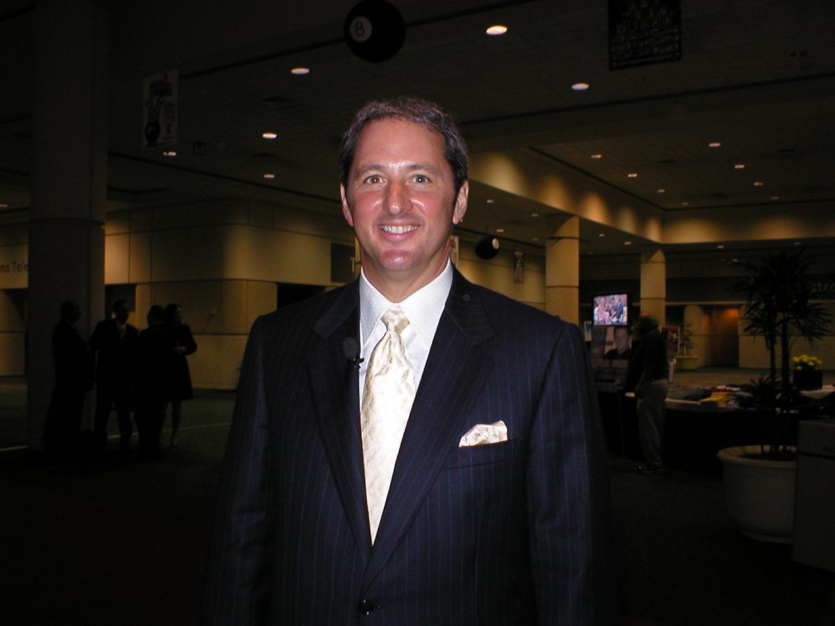 A waist-up photograph of a smiling man with short dark hair, wearing a pinstriped suit and gold tie, standing in a convention-center lobby.