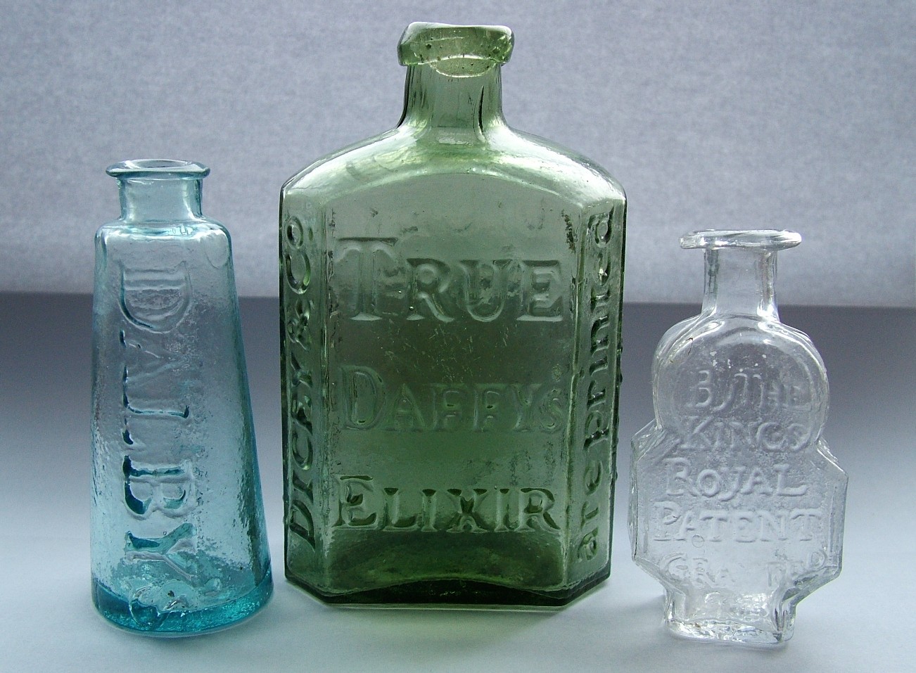 Photograph of three antique glass patent-medicine bottles standing side by side, with legible embossed lettering including 'DALBY,' 'TRUE DAFFYS ELIXIR,' and 'BY THE KINGS ROYAL PATENT GRANT[ED].'