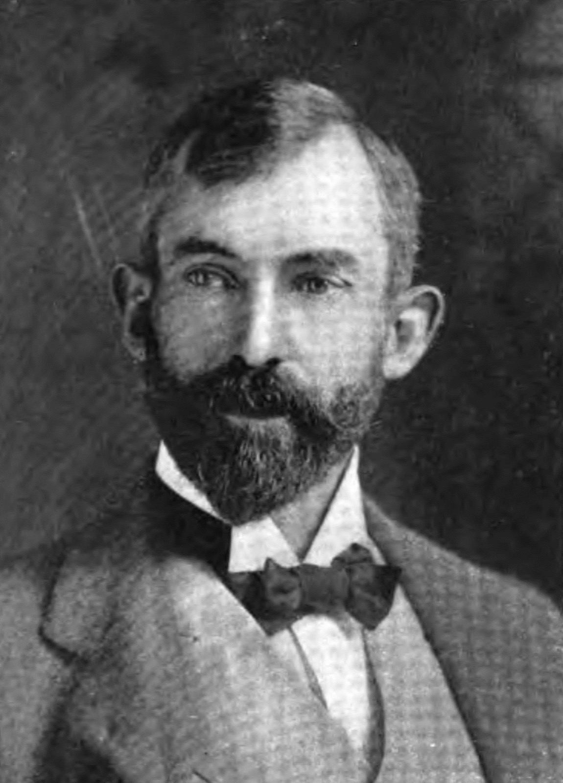 Black-and-white halftone portrait of a man with dark hair, a mustache, and a pointed beard, wearing a suit and bow tie; the dot pattern of magazine printing is visible.