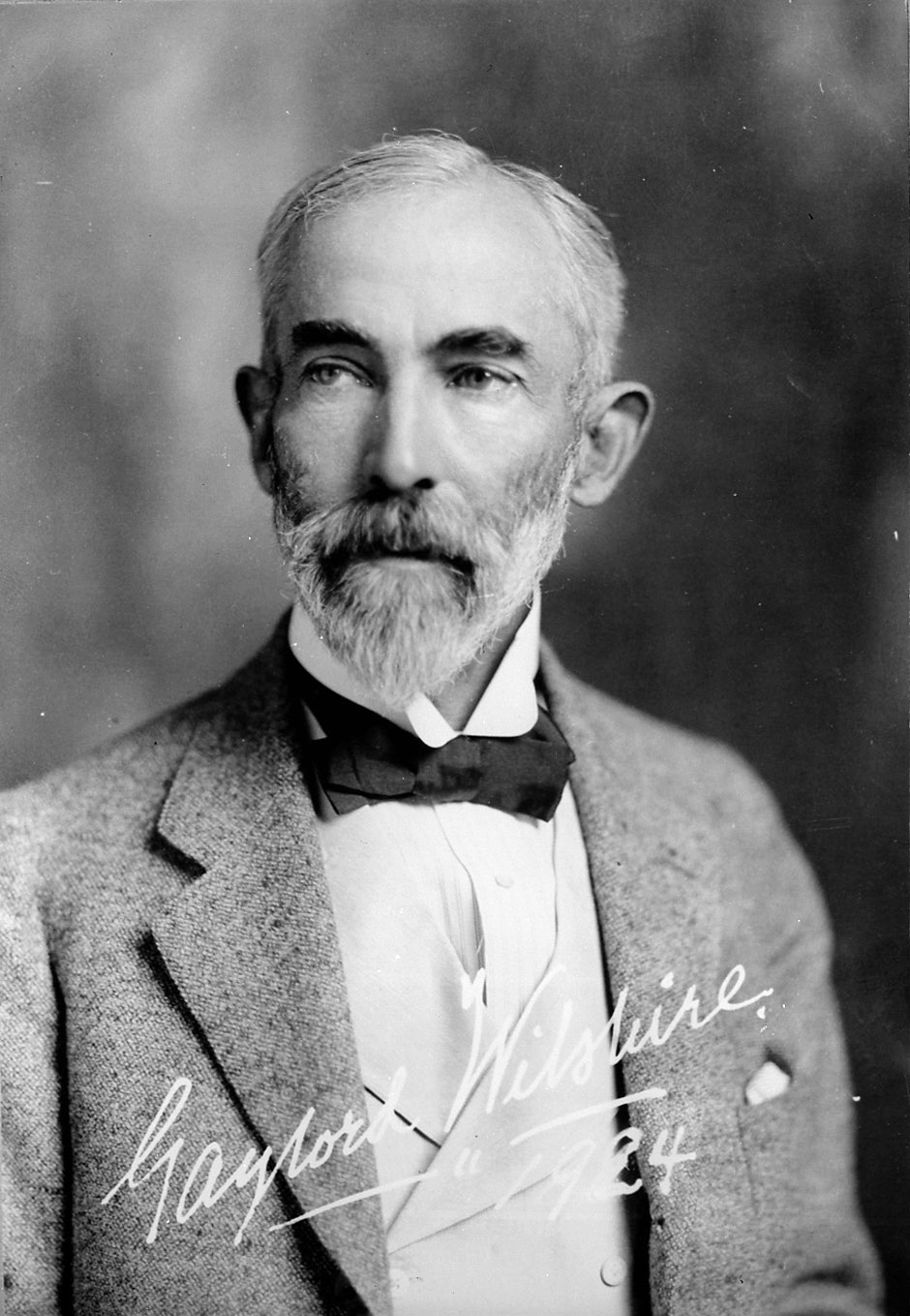 Black-and-white studio portrait of an older man with a full white beard, in a tweed jacket, wing collar, and bow tie; a handwritten signature reading 'Gaylord Wilshire 1924' runs across the lower part of the image.